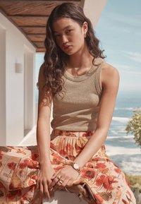 Beiges, geripptes Tanktop kombiniert mit einem fließenden, geblümten Rock in sanften Farbtönen. Accessoires umfassen eine silberne Uhr und goldene Ringe.