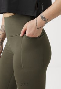 Olivengrønne høytalende leggings med synlig søm, sidelomme og logo detalj nederst. Elastisk, tettsittende materiale.