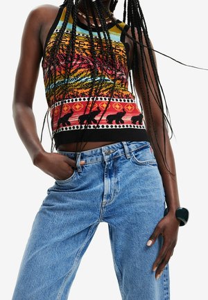 Woman wearing a multicolored sleeveless top with animal patterns and light blue high-waisted jeans, with one hand in pocket and braided hair.