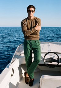 Homme portant des lunettes de soleil, un pull marron, un pantalon vert et des chaussures marron, debout les bras croisés sur un bateau blanc avec l'océan en arrière-plan.