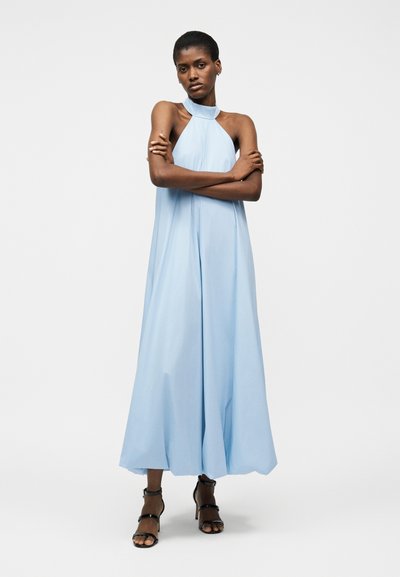 Femme aux cheveux courts portant une robe midi sans manches bleu clair à col halter et des sandales noires à talons hauts, debout les bras croisés.