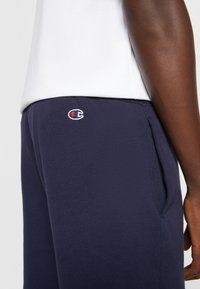 Tmavě modré bavlněné tepláky s bočními kapsami a malým vyšívaným logem na stehně. Hladká textura s elastickým pasem.