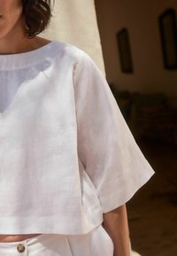 Bílé lněné krátké tričko s širokým výstřihem a krátkými rukávy. Lehká textura s jemným vzorem. V kombinaci se sladěnými vysokými kalhotami.