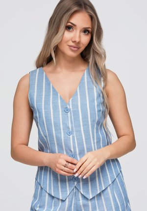 Giovane donna con lunghi capelli biondi indossa un top senza maniche a righe verticali blu e bianche con bottoni e un fondo abbinato, posando con le mani giunte.