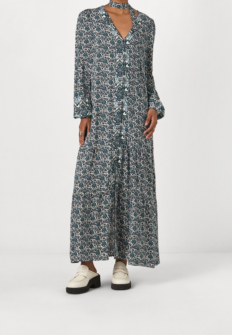 Femme portant une longue robe à motifs avec col en V et collier ras du cou assorti, associée à des mocassins blancs sur un fond uni.