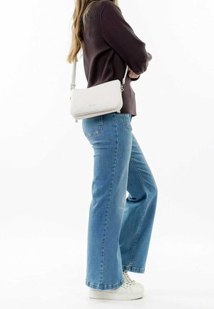 Kobieta ubrana w rozkloszowane niebieskie dżinsy, białe trampki i ciemny prążkowany sweter, niosąca małą białą torbę na ramię, stojąca bokiem.