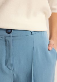 Jasnoniebieskie spodnie wykonane z gładkiego materiału, z przodem plisowanym, kieszeniami bocznymi i niebieskim guzikiem w talii; minimalistyczny design.