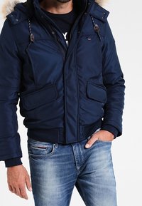 Man med en marinblå vadderad jacka med pälsbesatt huva och ljusblå urtvättade jeans, en hand i fickan.