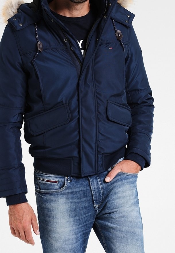 Man med en marinblå vadderad jacka med pälsbesatt huva och ljusblå urtvättade jeans, en hand i fickan.