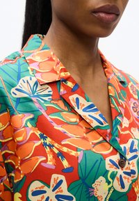 Γυναίκα που φοράει πολύχρωμο πουκάμισο με κουμπιά, διακοσμημένο με μεγάλα λουλουδένια και φυλλώδη σχέδια σε πορτοκαλί, πράσινο, μπλε και λευκό σε κόκκινο φόντο.