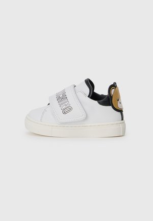 Baskets blanches en cuir avec des accents noirs, une bande velcro et un design d'ours au talon. Elles présentent une semelle texturée et des détails cousus.