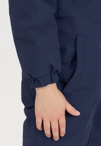 Marineblå vandtæt jumpsuit med elastiske manchetter, med "W-PRO 10000" prægning på ærmet og detail af sidelomme.