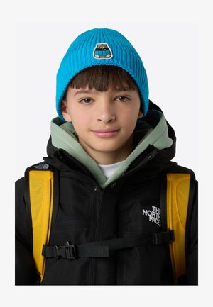 Gorro de punto acanalado azul con un parche de oso, llevado con una chaqueta negra y mochila amarilla. Destaca una sudadera verde con capucha debajo.
