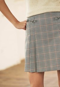 Jupe écossaise avec des motifs noirs, blancs et orange, un design plissé et des éléments décoratifs à la taille. Tissu léger.