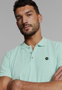 Mintgroene polo met een gestructureerde afwerking, uitgerust met een kraag met twee knopen en een hexagonaal logoaccent op de borst.