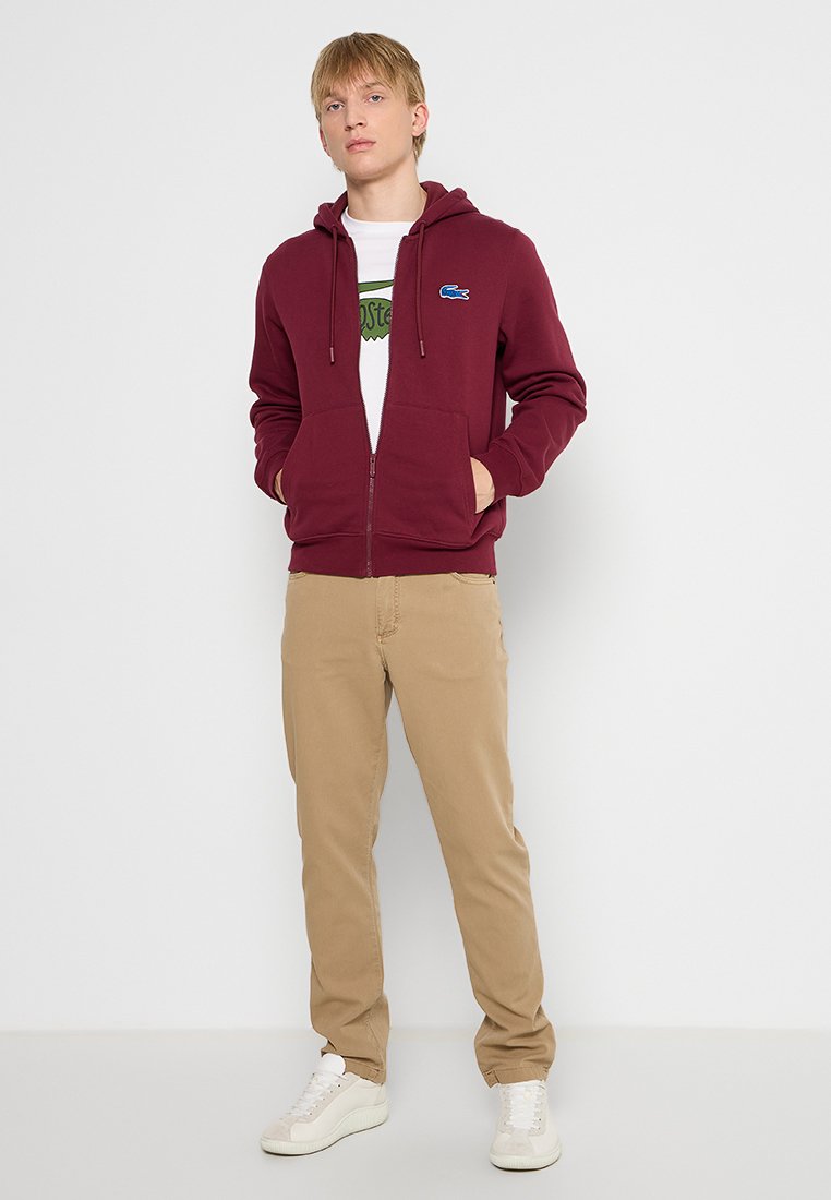 Haut à capuche zippé bordeaux avec poches kangourou, poignets côtelés et un logo bleu. Porté avec un pantalon beige et des baskets blanches. Texture de tissu lisse.