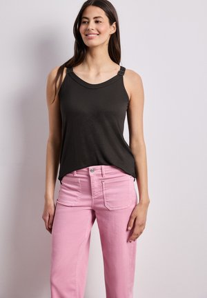 Vrouw draagt een zwarte mouwloze top met gespen aan de bandjes en roze jeans met hoge taille, staand tegen een effen lichte achtergrond.