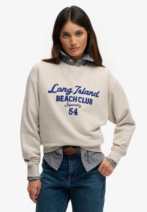 Vrouw met een beige sweatshirt met blauwe tekst "Long Island Beach Club Superdry 54", gecombineerd met een gestreept overhemd en een blauwe spijkerbroek met een bruine riem.