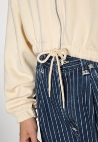 Beige mikina na zip s funkční šňůrkou uvázanou přes modré džíny s bílými vertikálními pruhy a distressed detaily.