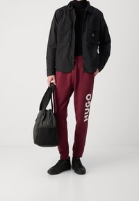 Schwarze Zip-up-Jacke mit Taschen, eng anliegender schwarzer Rollkragenpullover, burgunderfarbene Sweatpants mit "HUGO"-Aufdruck und eine schwarze Sporttasche. Schwarze Sneakers.