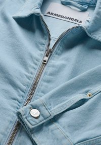 Hellblaue Jeansjacke mit silbernem Reißverschluss, umgeschlagenem Kragen und Druckknopfdetails an der Manschette. Aus einem weichen, strukturierten Stoff gefertigt.