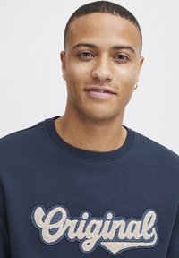 Tumši zilā džemperī ar ribu apkakli ir tekstūras krēmbalts "Original" izšūts uzlīme uz krūtīm.