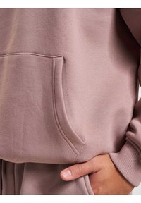 Lys mauve sweatshirt med kængurulomme. Blød tekstur, ribbede manchetter og synlige syninger. En hånd hviler i lommen.