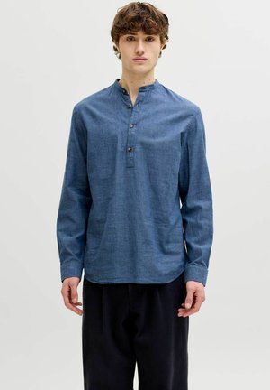 Joven con una camisa henley azul de manga larga con botones de madera y pantalones negros, de pie frente a un fondo claro y liso.