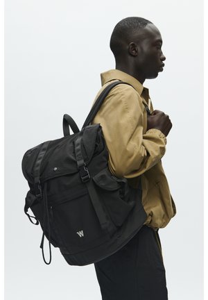 Sac à dos en tissu noir avec des bretelles réglables, une fermeture à rabat, une poche avant et un logo argenté. Le modèle porte une veste beige.