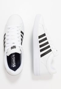 White sneakers with a smooth design featuring three black stripes, lace closure, and a textured white rubber sole. Notable brand logo inside.