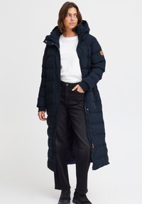 Lang, marineblå puffjakke med hette, kviltet design, svarte ribbestrikkede mansjetter, glidelås foran og trykknapper. Brukt over en hvit skjorte og svarte jeans.