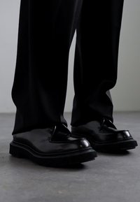 Chaussures en cuir noir à finition lisse, avec un bout arrondi et une semelle épaisse et texturée. Portées avec un pantalon noir, sur fond neutre.