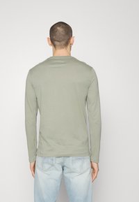 Mann mit kurzem Haar, der ein hellgrünes Langarmshirt und hellblaue Jeans trägt, steht vor einem schlichten weißen Hintergrund.