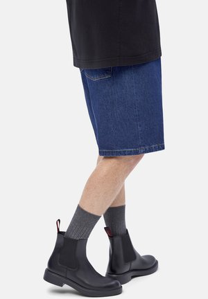 Persona con calcetines grises hasta la mitad de la pantorrilla, botas de cuero negro hasta el tobillo y pantalones cortos de mezclilla azules, de pie contra un fondo liso.