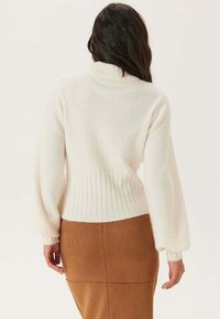 Pull blanc et moelleux avec un col montant, taille et poignets côtelés, associé à une jupe marron jusqu'au genou. Texture douce, coupe décontractée.