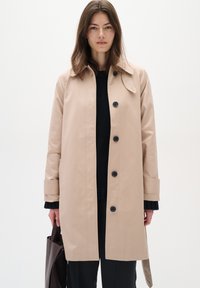 Beige trenčkot vyrobený z hladkého materiálu, s klasickým límcem, šesti černými knoflíky a bočními kapsami, kombinovaný s černým pleteným topem.