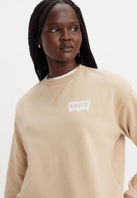 Sudadera beige de Levi's con un logo blanco, con un cuello redondo y tejido texturizado. Modelo posando con trenzas frente a un fondo liso.