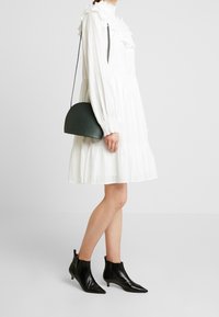 Vit klänning med volangkrage och ärmar, lager-på-lager kjol, kombinerad med en mörkgrön rund handväska och svarta spetsiga ankelstövlar.