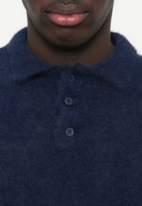 Marineblaues gestricktes Poloshirt mit einer flauschigen Textur, das einen Kragen, drei Knöpfe und ein dezentes Logo-Detail am Saum aufweist.
