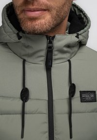 Chaqueta de plumas verde con cierre de cremallera, acentos negros y cordones; presenta un parche de marca y un diseño con capucha con una textura suave.