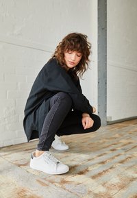 Chemise noire oversize, jean gris avec surpiqûres blanches, et baskets blanches épaisses avec accents gris, posés sur un sol en bois.