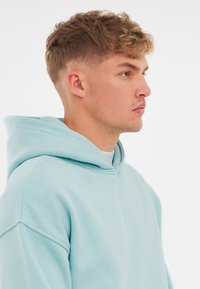 Felpa blu chiaro con un fit rilassato, ampio cappuccio e tessuto texturizzato. Il design include dettagli di cucitura sottili sulle spalle.