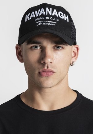 Mladý muž so sivými očami, ktorý nosí čiernu čiapku s textom "Kavanagh Winners Club" a čierne tričko, otočený čelom k hľadisku na jednoliatom pozadí.