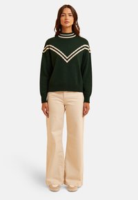 Maglione in maglia verde scuro con colletto alto, caratterizzato da strisce a zig zag bianche. Abbinato a pantaloni a gamba larga beige chiaro e scarpe con zeppa.
