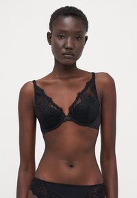 Reggiseno in pizzo nero con ricami floreali, coppe sagomate e spalline regolabili. Il tessuto ha una consistenza liscia con bordi ondulati.