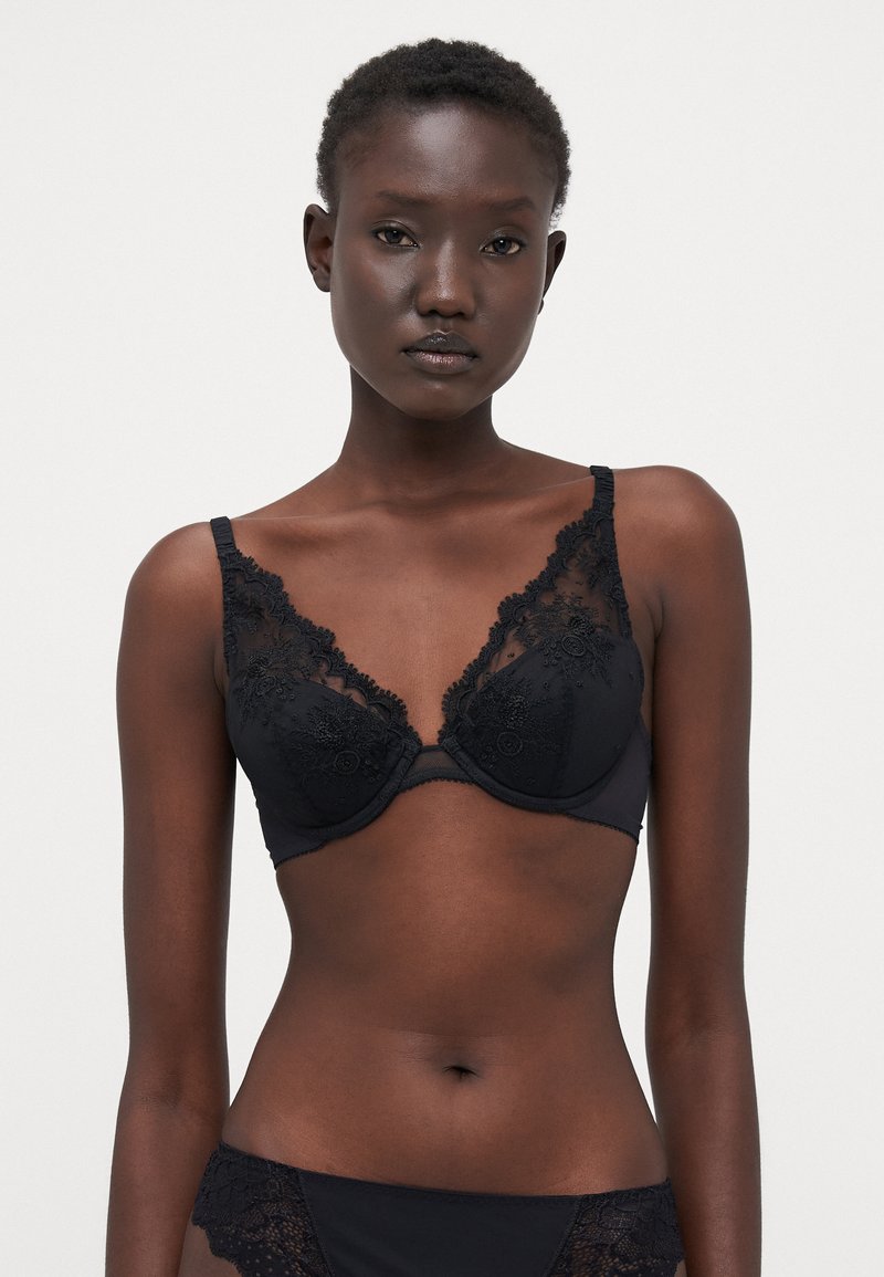Reggiseno in pizzo nero con ricami floreali, coppe sagomate e spalline regolabili. Il tessuto ha una consistenza liscia con bordi ondulati.