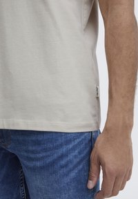 Beige bomull t-skjorte med rett skjæring i bunnen, søm detaljer, og et stoffmerke på siden. Brukt sammen med blå denim jeans.