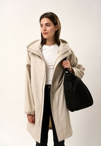Cappotto impermeabile beige con cappuccio, caratterizzato da una linea dritta e zip frontale. Abbinato a una borsa nera con cordino.