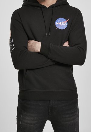 Osoba ubrana w czarną bluzę z kapturem NASA, z założonymi na piersi rękami, logo NASA na klatce piersiowej i naszywkami na lewym rękawie, w połączeniu z ciemnymi dżinsami.