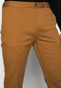 Hnědé bavlněné chino kalhoty s hladkou texturou, vybavené zapínáním na knoflíky, poutky na opasek a předními kapsami.